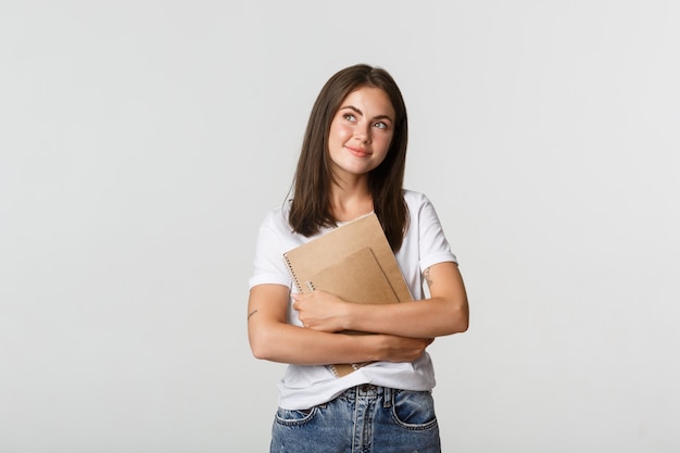 Portrait d'étudiante nostalgique souriante à la recherche dans le coin supérieur gauche, tenant des cahiers.