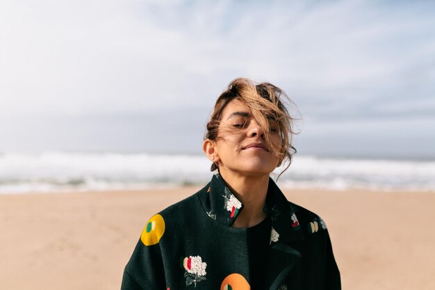 Portrait extérieur d'une femme romantique aux cheveux volants se tient sur la plage de sable avec les yeux fermés et un sourire heureux sur fond de ciel bleu