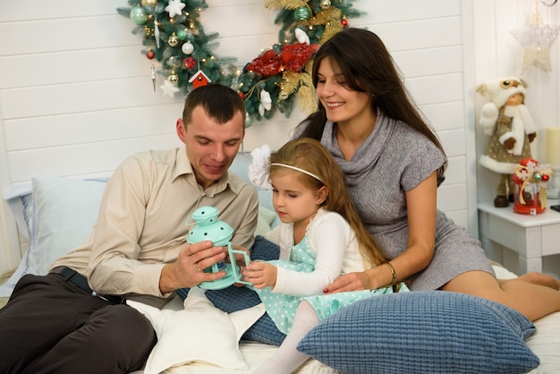 Portrait de famille heureuse à noël, mère, père et enfant assis sur le lit et allumant une bougie à la maison, décoration de noël autour d'eux