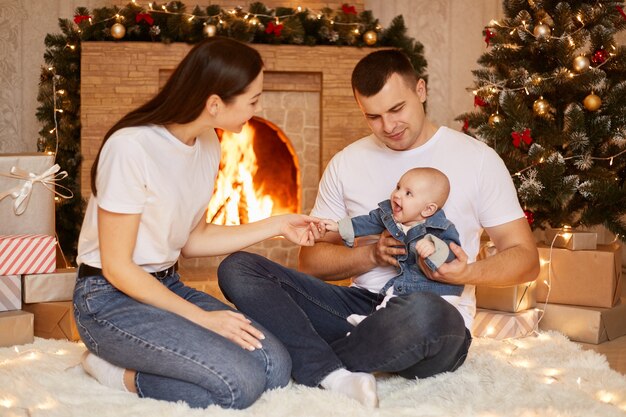 Portrait d'une famille heureuse, d'un père, d'une mère et d'une fille en bas âge posant devant une cheminée et un arbre de Noël à la maison, heureux de célébrer les vacances d'hiver ensemble, bonne année.