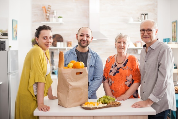 Portrait de famille multigénération souriante à la caméra dans la cuisine avec paparbag d'épicerie et une variété de fromages