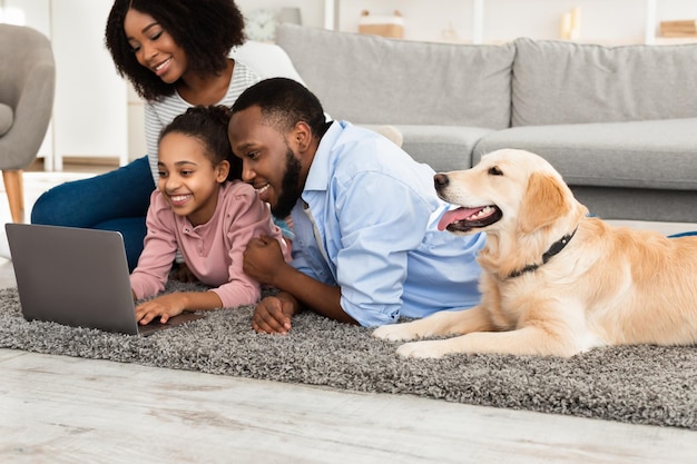 Portrait d'une famille noire heureuse et aimante allongée sur un tapis de sol avec leur chien domestique, utilisant un ordinateur portable et regardant des vidéos ou des films, naviguant sur internet, passant du temps ensemble à la maison dans le salon