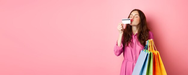 Portrait d'une femme accro du shopping heureuse et excitée tenant des sacs à provisions et embrassant une carte de crédit en plastique smi