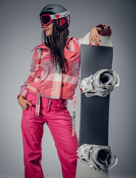 Portrait de femme active dans un costume de ski rose tenant un snowboard. Isolé sur fond gris.