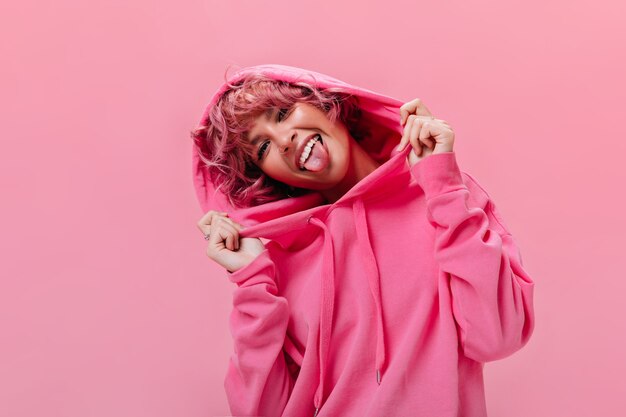 Portrait d'une femme active et joyeuse aux cheveux roses en sweat à capuche surdimensionné fuchsia montre la langue et fait une grimace sur un mur isolé