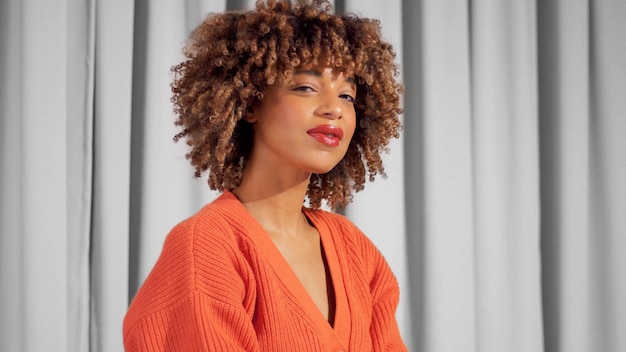 Portrait de femme adulte noire de race mixte sur des rideaux gris en veste tricotée orange vif et maquillage naturel