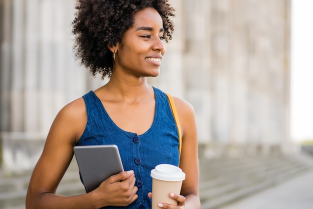 Portrait de femme d'affaires afro tenant une tablette numérique et une tasse de café en se tenant debout à l'extérieur dans la rue. Concept d'entreprise et urbain.