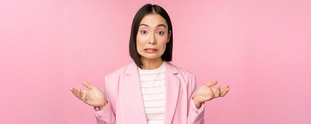 Portrait d'une femme d'affaires asiatique confuse, haussant les épaules, l'air désemparé et perplexe, je ne sais pas dire debout sur fond rose en costume de bureau