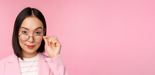 Portrait d'une femme d'affaires asiatique dans des verres regardant intriguée par la caméra et une vendeuse professionnelle souriante regardant avec intérêt sur fond rose