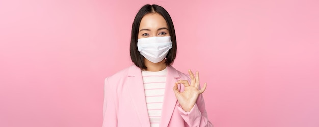 Portrait d'une femme d'affaires asiatique portant un masque médical montrant un signe correct portant des règles de travail en costume pendant la pandémie de covid19 debout sur fond rose