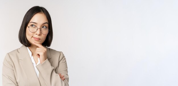 Portrait d'une femme d'affaires asiatique réfléchie dans des verres faisant des suppositions pensant debout en costume beige sur fond blanc