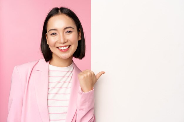 Portrait d'une femme d'affaires asiatique souriante en costume dame d'entreprise pointant le doigt sur un panneau mural blanc vide avec des informations ou une publicité debout sur fond rose