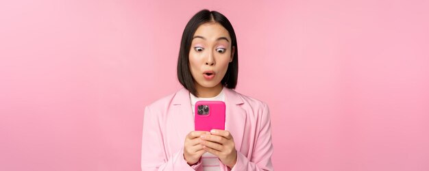 Portrait d'une femme d'affaires asiatique avec un visage surpris à l'aide d'une application pour smartphone portant un costume d'affaires Fille coréenne avec un téléphone portable et une expression de visage excité sur fond rose