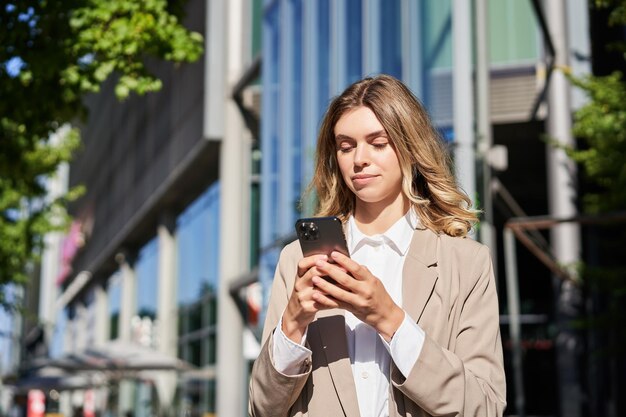 Portrait d'une femme d'affaires confiante debout dans la rue près d'un immeuble de bureaux à l'aide d'une commande de téléphone portable