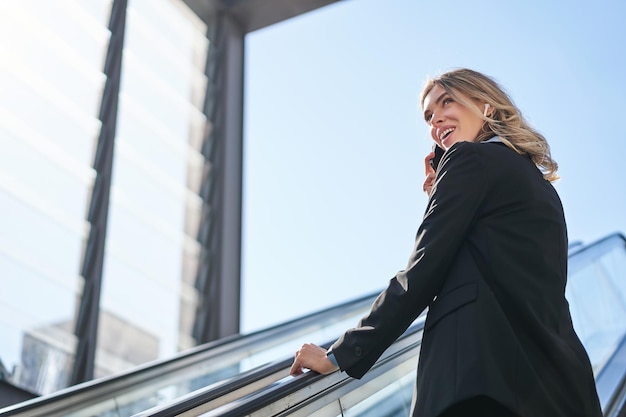 Photo gratuite portrait de femme d'affaires en costume noir qui monte sur l'escalator en parlant sur la vendeuse de téléphone mobile wal