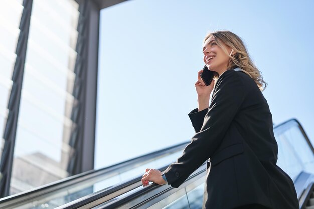 Portrait de femme d'affaires en costume noir qui monte sur l'escalator en parlant sur la vendeuse de téléphone mobile wal