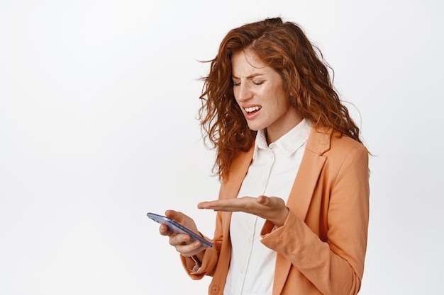 Portrait d'une femme d'affaires regardant fixement confus un téléphone portable se disputant à propos de quelque chose sur un smartphone debout sur fond blanc