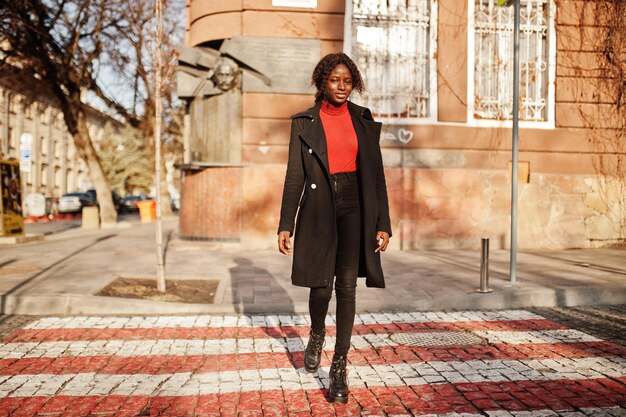 Portrait d'une femme africaine aux cheveux bouclés portant un manteau noir à la mode et un col roulé rouge marchant sur le passage clouté