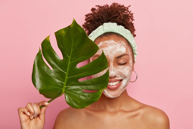 Portrait d'une femme afro-américaine heureuse couvre la moitié du visage avec des feuilles vertes, nettoie le visage, se lave avec du savon à bulles, se tient topless, se soucie de sa beauté et de son corps