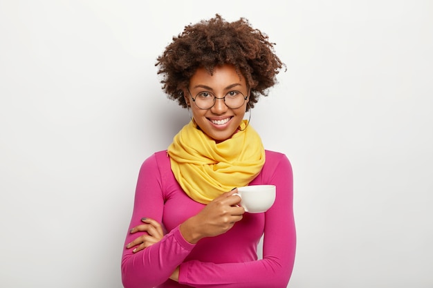 Portrait De Femme Afro-américaine Joyeuse Avec Une Expression Heureuse, Porte Des Lunettes Optiques, Détient Une Tasse De Boisson, Porte Des Lunettes Optiques, Col Roulé Rose Et écharpe, Isolé Sur Fond Blanc