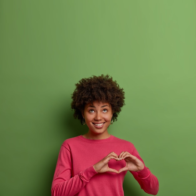 Portrait de femme afro-américaine à la recherche agréable et joyeuse montre signe de coeur sur la poitrine, exprime l'amour et les soins, regarde au-dessus, porte un pull rose, pose contre le mur vert, l'espace vide vers le haut