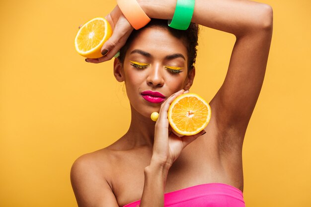 Portrait de femme afro-américaine sensuelle avec les yeux fermés tenant deux parties d'orange à la caméra en appréciant les agrumes isolés, sur le mur jaune