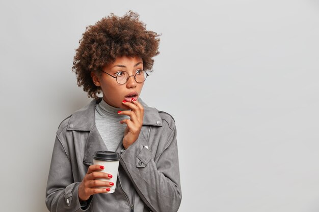 Portrait de femme afro-américaine surpris regarde choqué côté boissons café à emporter porte des lunettes rondes veste à la mode isolé sur mur gris