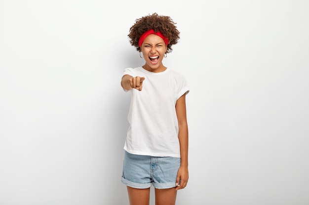 Portrait d'une femme afro ravie de rire de quelque chose de drôle, pointe directement dans la caméra, exprime de bonnes émotions, porte un bandeau rouge, un t-shirt et un short, des modèles sur un mur blanc.