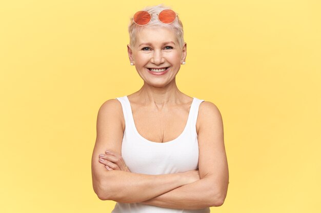 Portrait de femme d'âge moyen confiant avec corps sportif bronzé bénéficiant d'un temps chaud et ensoleillé regardant la caméra avec un sourire heureux, croisant les bras sur sa poitrine, portant débardeur blanc et lunettes