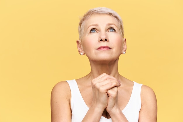 Portrait de femme d'âge moyen nerveuse frustrée avec de courts cheveux blonds levant les mains et tenant les mains jointes, ayant une expression faciale pleine d'espoir, priant Dieu, demandant de l'aider à traverser les moments difficiles