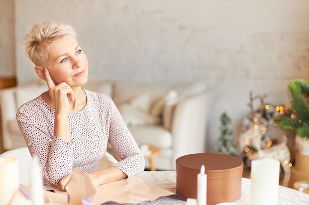 Portrait de femme d'âge moyen réfléchie en robe élégante assis à table dans la chambre décorée avec une guirlande ayant un regard pensif, tenant l'index sur sa tête, pensant comment emballer des cadeaux pour la famille