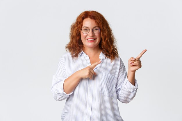 Portrait d'une femme d'âge moyen souriante agréable aux cheveux rouges, portant des lunettes et un chemisier montrant la publicité, le client de l'entreprise recommande un produit ou un service, pointant vers la droite.