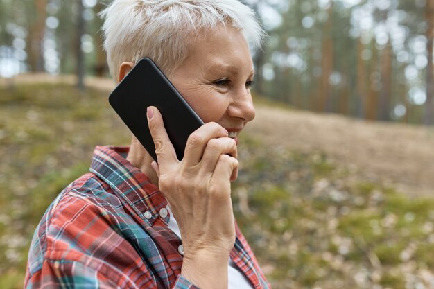 Portrait de femme d'âge moyen avec wrikles posant à l'extérieur en chemise à carreaux tenant un téléphone intelligent à son oreille, ayant une belle conversation, profitant d'une promenade en forêt.