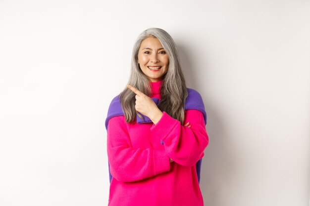 Portrait d'une femme âgée asiatique heureuse souriante, pointant vers le coin supérieur gauche et semblant satisfaite, démontre une promotion spéciale, debout sur fond blanc.