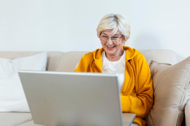 Portrait d'une femme âgée assise à la maison sur le canapé et travaillant sur son ordinateur portable