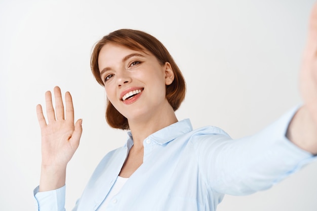 Photo gratuite portrait d'une femme agitant la main pour dire bonjour sur un chat vidéo, tenant un smartphone dans la main tendue, saluant un ami, debout contre un mur blanc