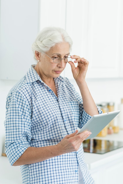 Portrait, femme aînée, debout, cuisine, regarder, tablette numérique