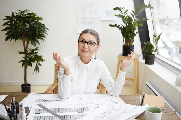 Portrait de femme architecte senior d'âge moyen expérimenté dans des lunettes élégantes et chemisier blanc assis sur son lieu de travail avec des dessins de projet architectural sur le bureau, se réjouissant du travail bien fait