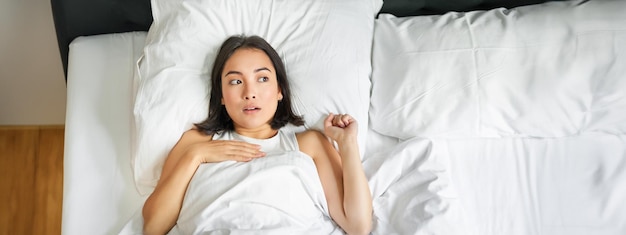 Portrait d'une femme asiatique allongée dans son lit avec un visage choqué, l'air surprise et bouleversée, haletant de qch