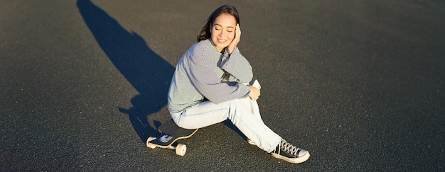 Photo gratuite portrait d'une femme asiatique assise sur un skateboard patinant sur son longboard de croiseur en utilisant une application pour smartphone