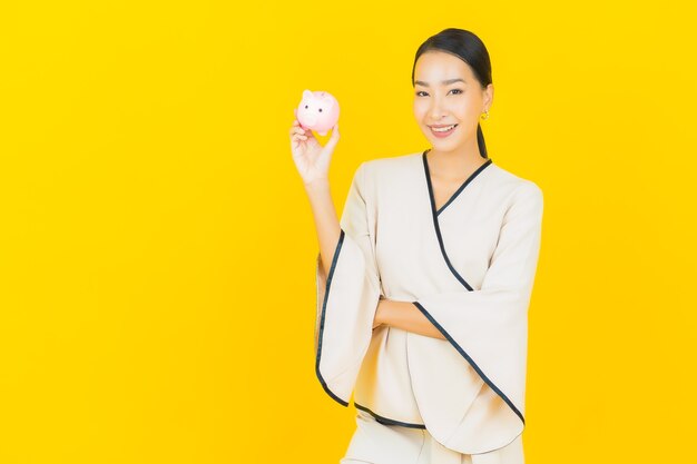 Portrait de femme asiatique belle jeune entreprise avec beaucoup d'argent comptant et tirelire sur mur jaune