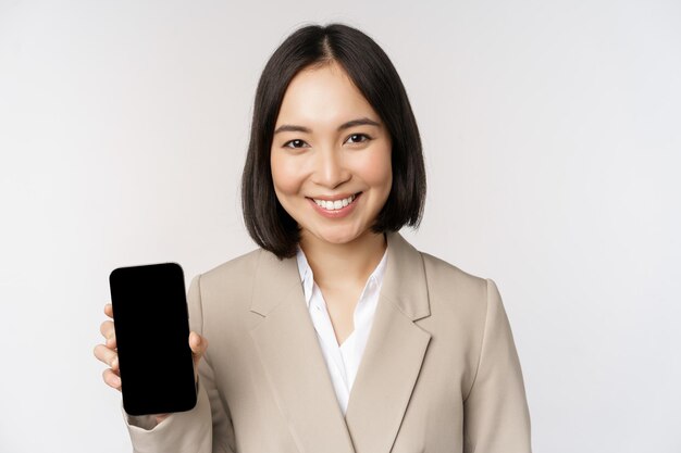 Portrait de femme asiatique d'entreprise montrant l'écran du téléphone mobile de l'interface de l'application smartphone debout sur fond blanc