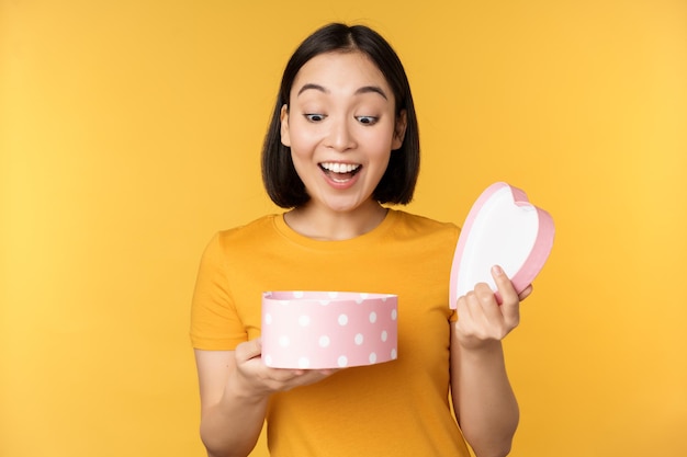 Portrait d'une femme asiatique excitée boîte-cadeau ouverte avec un visage heureux surpris debout sur fond jaune