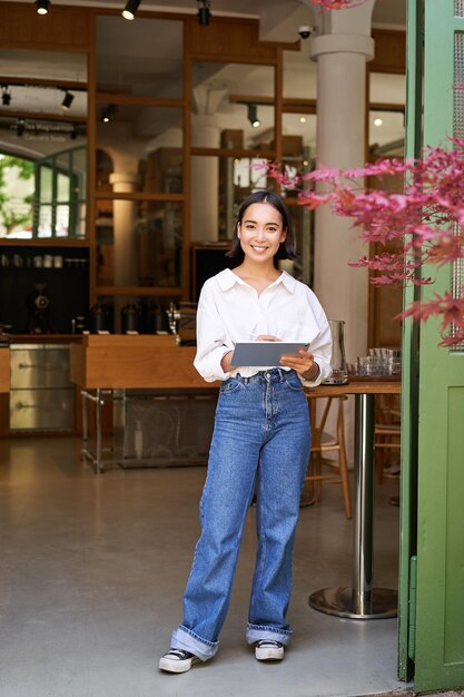 Photo gratuite portrait d'une femme asiatique gérante debout avec une tablette devant l'entrée du café accueille les invités