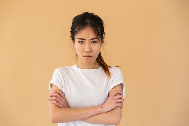 Portrait d'une femme asiatique indignée portant un t-shirt basique posant à l'avant avec un regard en colère isolé sur un mur beige en studio