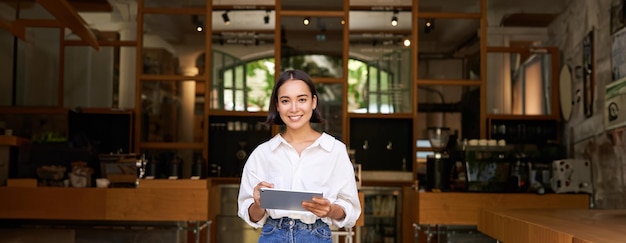 Photo gratuite portrait d'une femme asiatique manager debout avec une tablette devant l'entrée du café accueille les invités