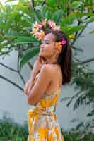 Photo gratuite portrait de femme asiatique en robe d'été jaune se dresse avec fleur thaï plumeria dans les cheveux et boucles d'oreilles rondes femme avec lumière composent à l'extérieur sur fond de mur et de buissons verts