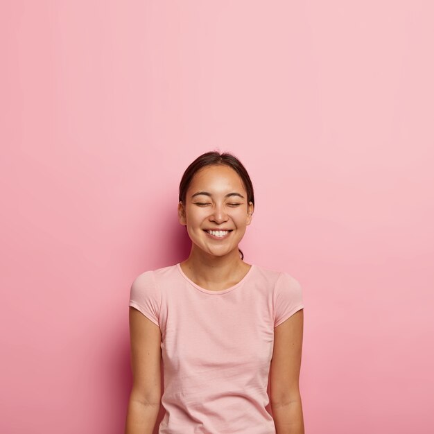 Portrait de femme asiatique satisfaite d'emotiove avec une beauté naturelle, cheveux peignés foncés, sourit joyeusement, garde les yeux fermés, porte un t-shirt décontracté, isolé sur un mur rose. Personnes, appartenance ethnique, émotions positives