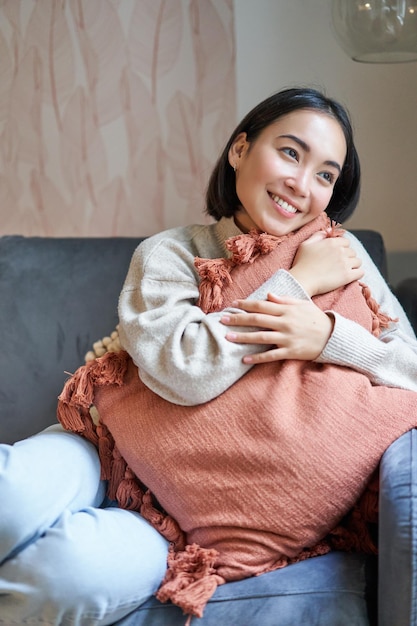 Photo gratuite portrait d'une femme asiatique se sentant à l'aise dans sa maison étreignant un oreiller sur un canapé et regardant un smili pensif