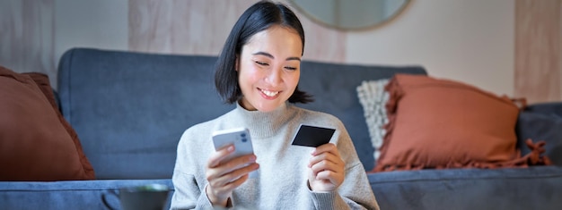 Photo gratuite portrait d'une femme asiatique souriante payant par carte de crédit sur son application pour smartphone organiser un prélèvement automatique